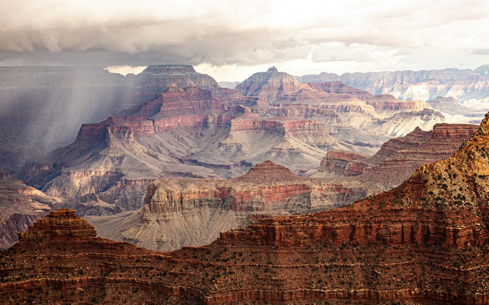 Layered red and cream canyon walls under a heavy cloud bank, with a shaft of rain falling on the left