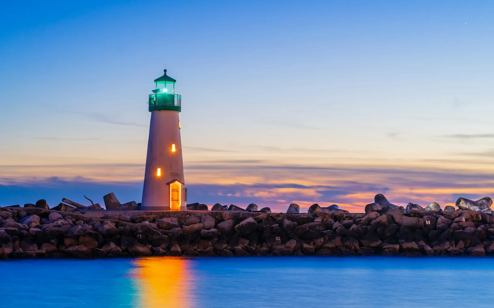 A white lighthouse with a glowing green lantern on a stone breakwater at dusk, its light reflected in the blue water