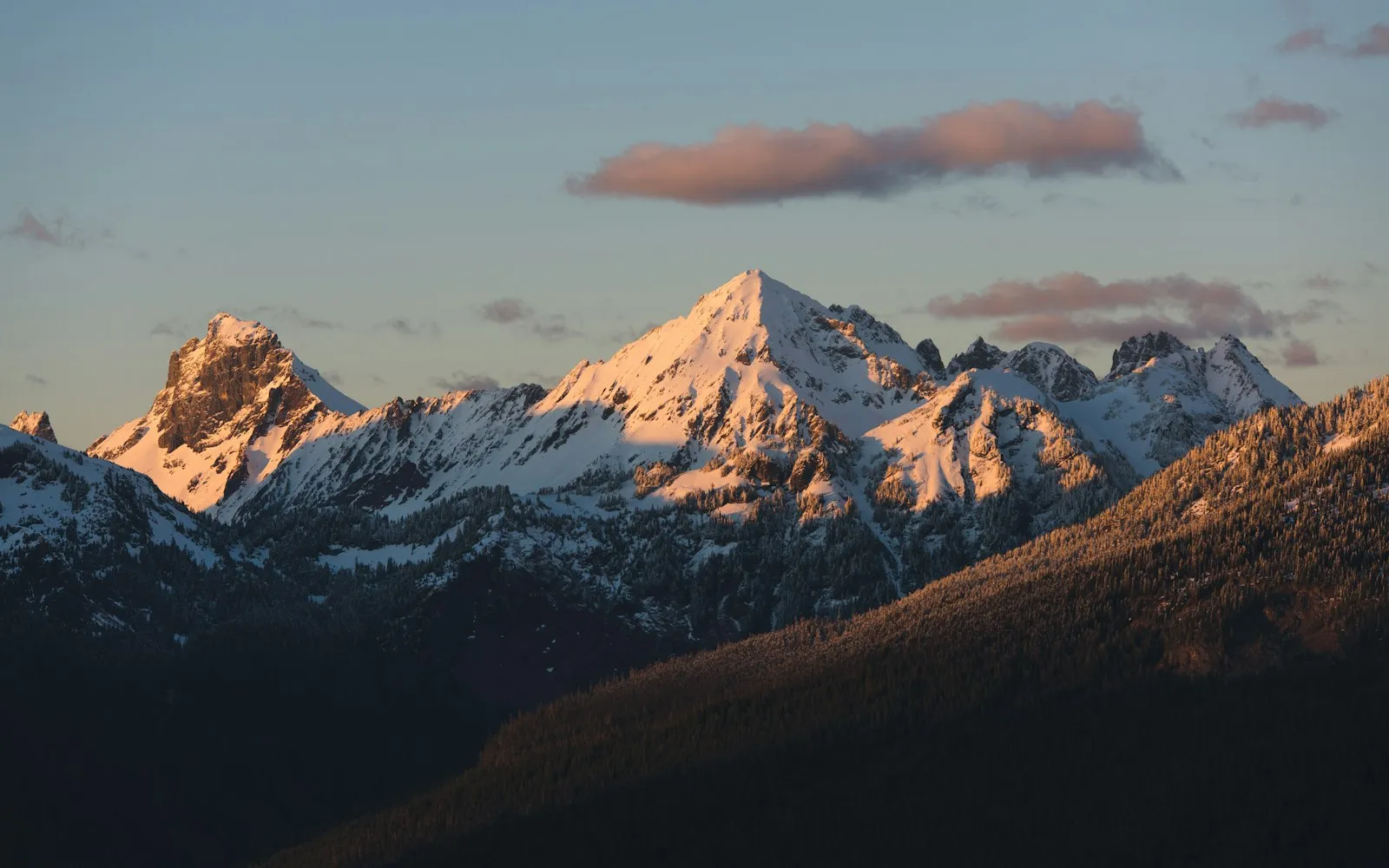 Low sun lighting snow-covered peaks in pink and gold above a dark forested slope