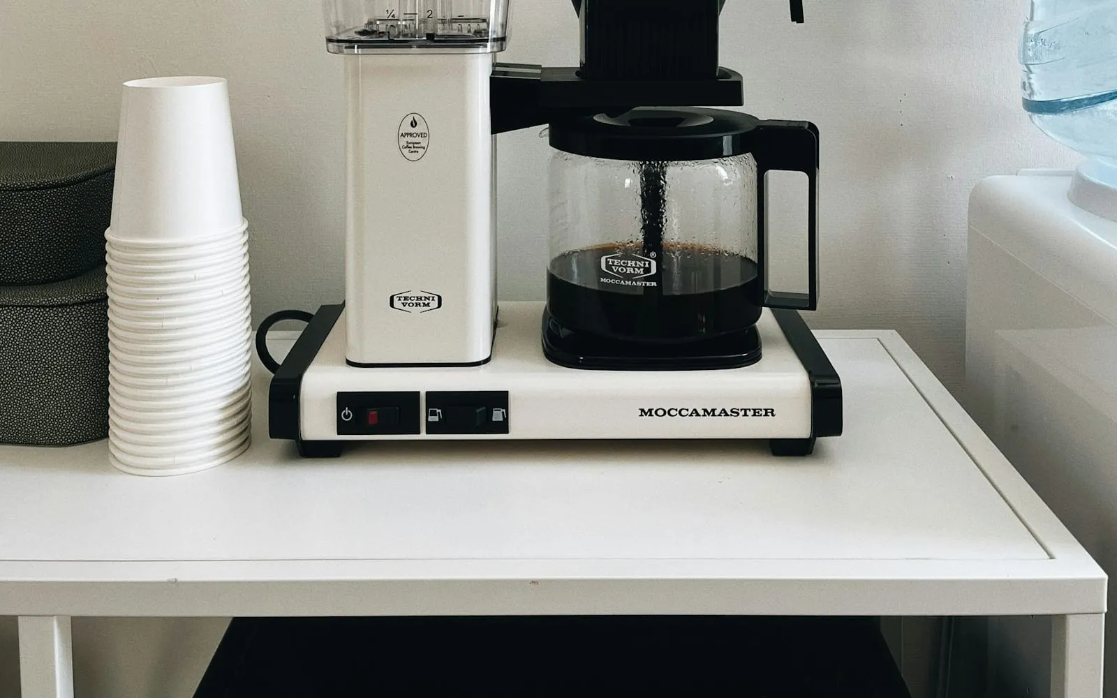 A filter coffee maker and a stack of paper cups on a white table