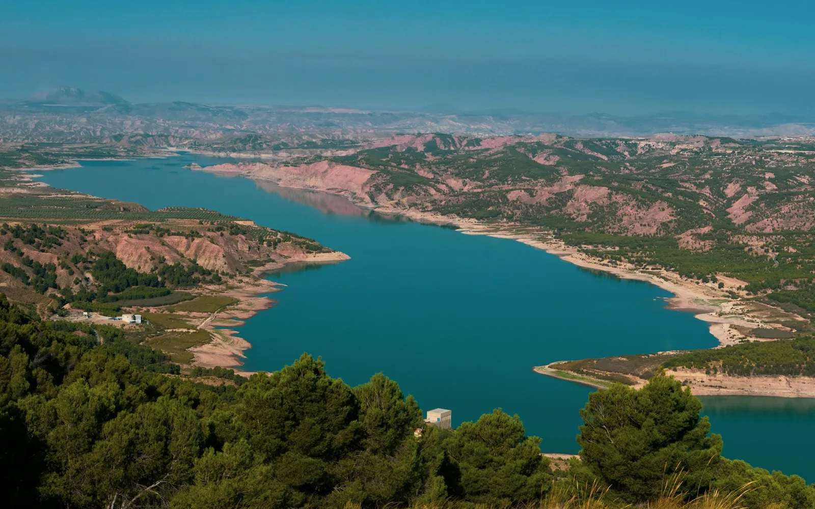 A winding turquoise reservoir among pine-covered hills and pale eroded slopes under a hazy blue sky