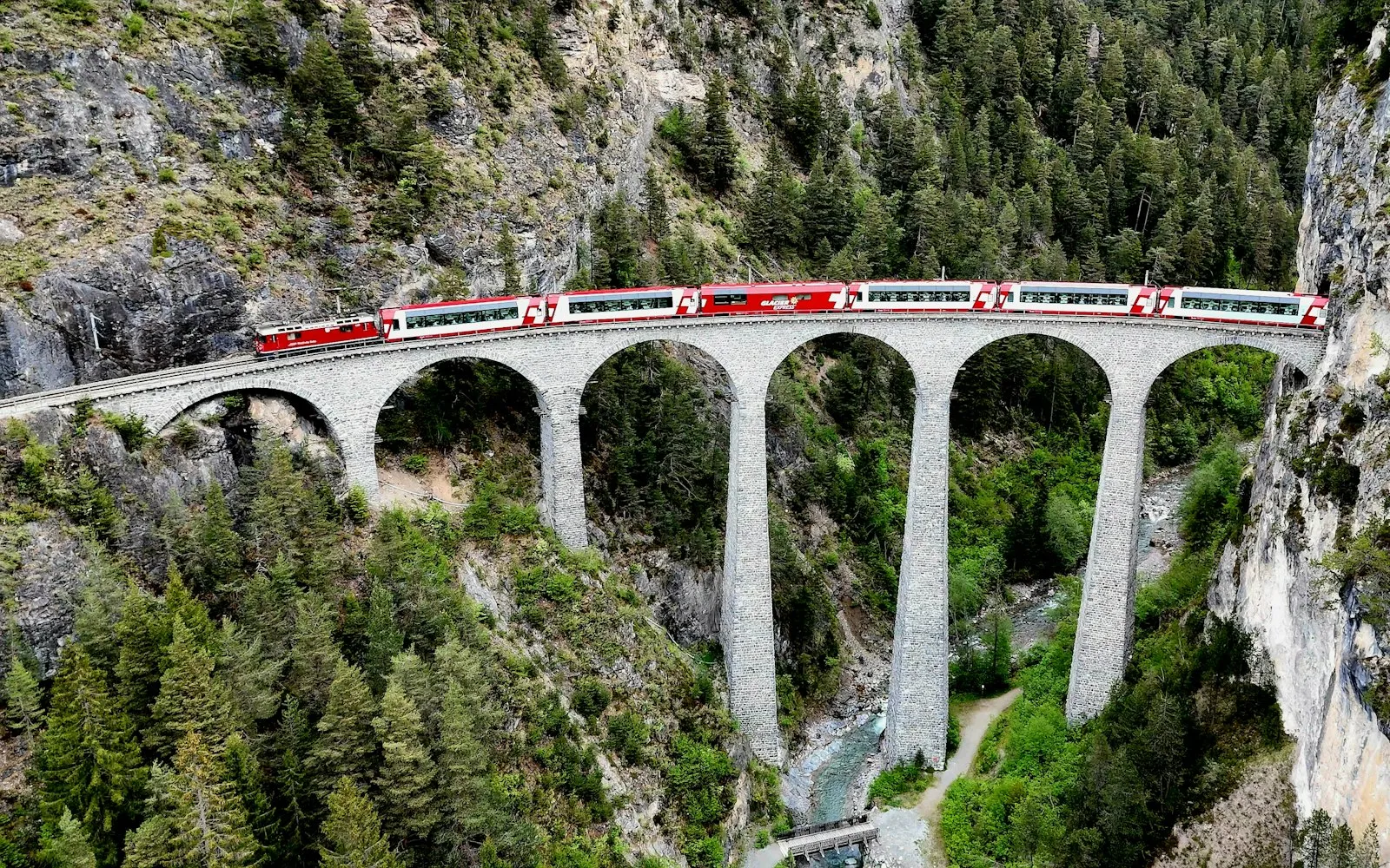 A red passenger train crossing a tall arched stone viaduct over a forested gorge