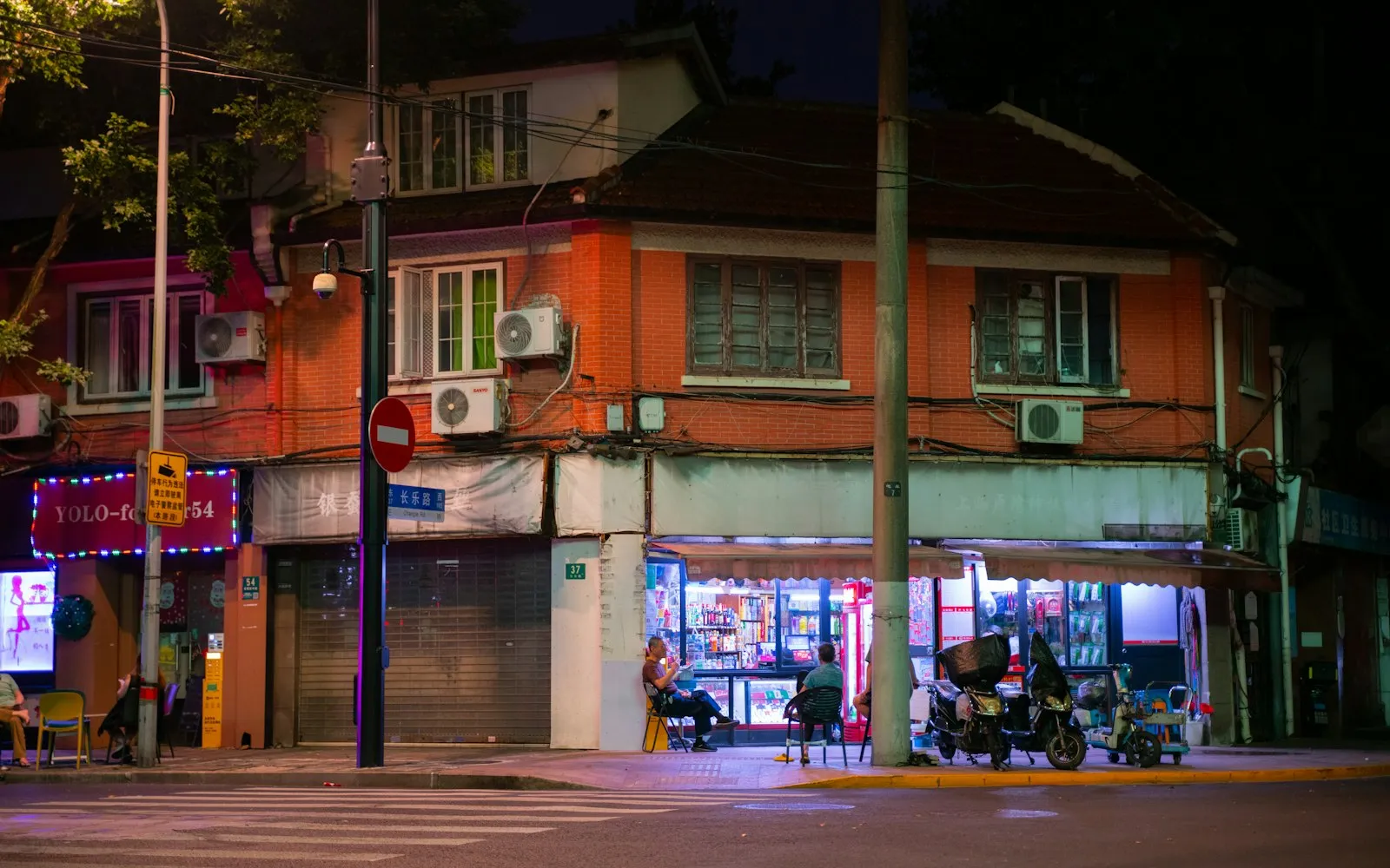 A lit corner shop at night, with people sitting outside under its awning