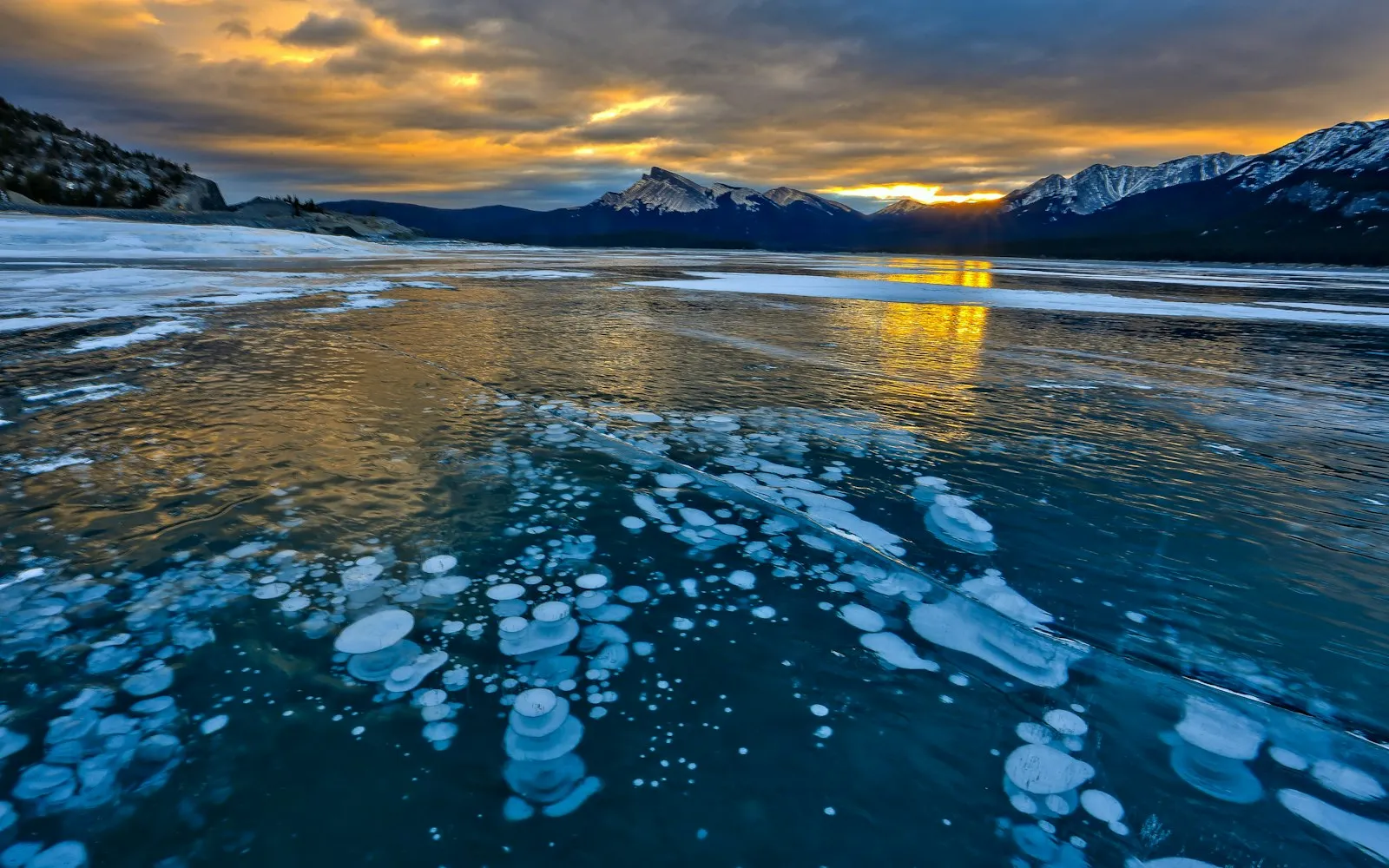 White bubbles frozen in clear blue lake ice, with snowy peaks and a golden sunset beyond