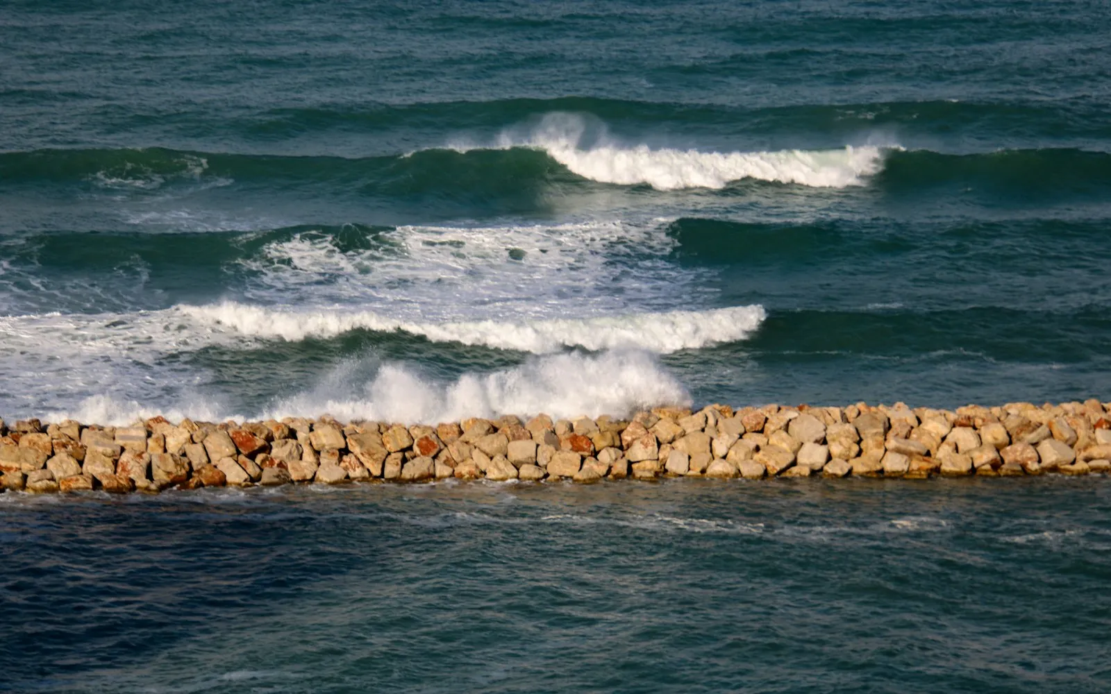 Rows of waves breaking against a long rock breakwater, with calmer water on the near side
