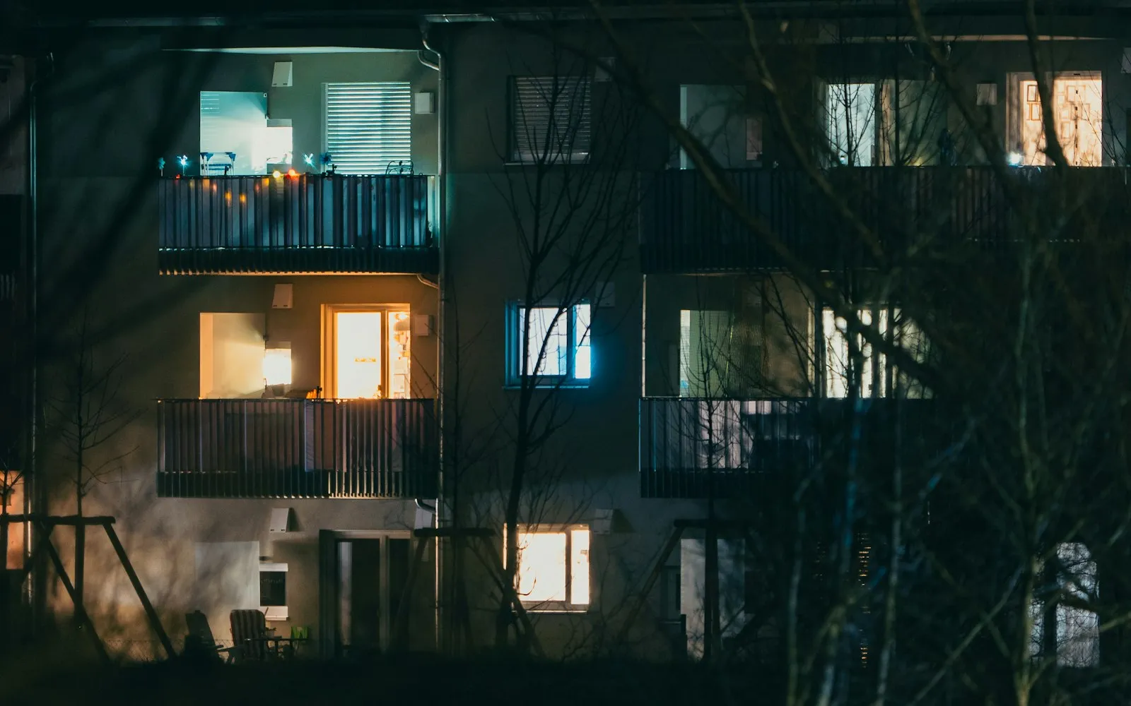 An apartment block at night with a few lit windows and balconies behind bare trees