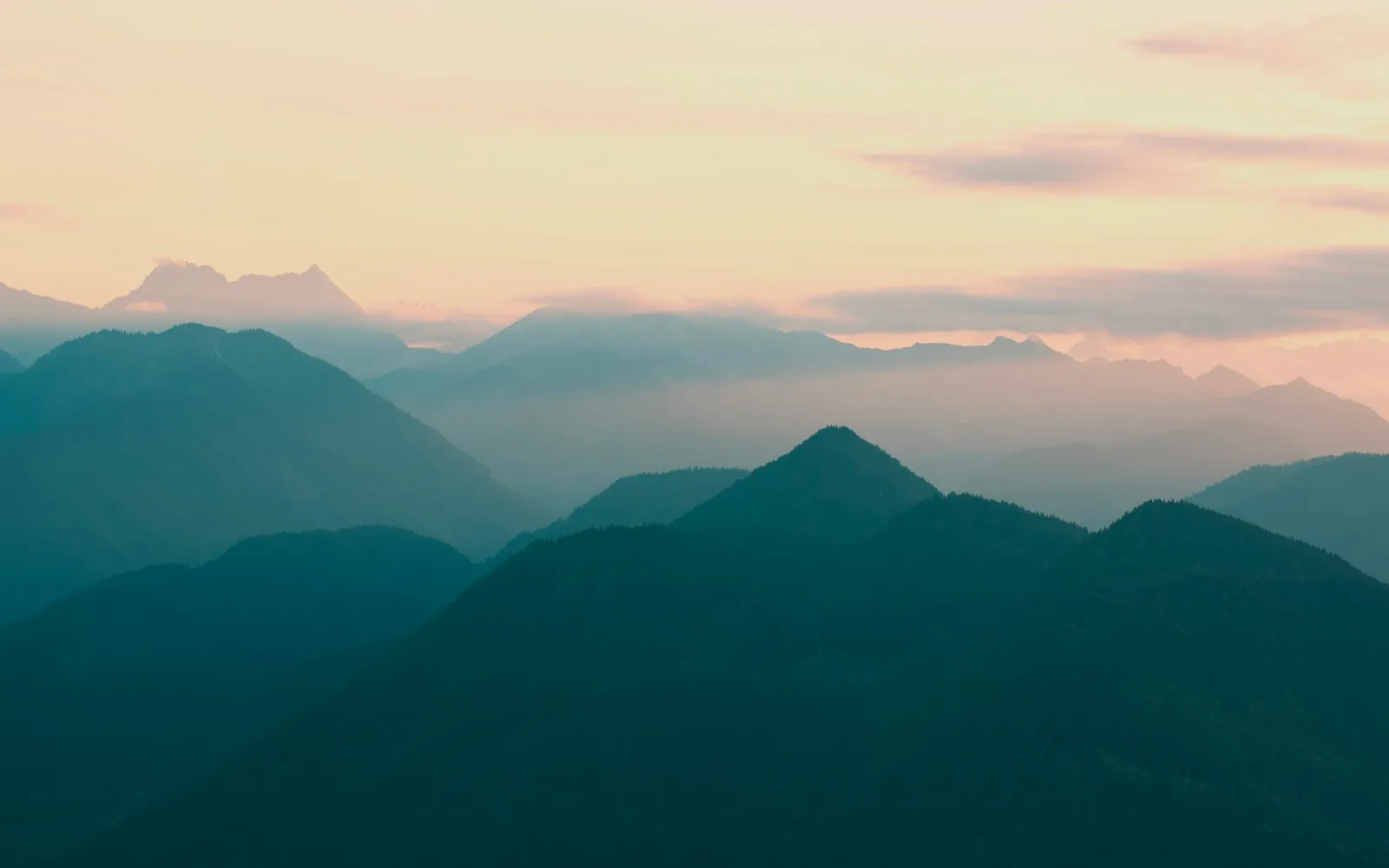 Layered forested mountain ridges fading into haze beneath a pale peach evening sky